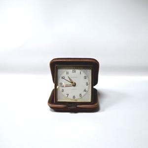 May include: A vintage square travel clock with a white face, gold hands, and gold numerals. The clock is housed in a brown leather case that opens to reveal the clock face. The clock face reads "Longines" and "Quartz".