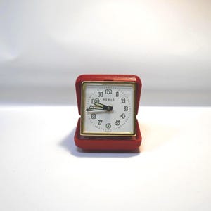 May include: A small, red travel alarm clock with a square face and gold-colored trim. The clock face is white with black numerals and hands. The brand name "REBUS" is printed on the face. The clock is in an open, red case.