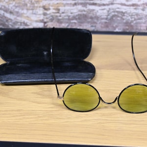 May include: A pair of round, black framed sunglasses with yellow lenses. The sunglasses are sitting on a wooden surface next to a black leather case.