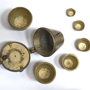 Può includere: Un assortimento di misurini antichi in ottone e un contenitore incernierato, tutti con una patina invecchiata. La collezione comprende varie dimensioni e forme, suggerendo un set per misurazioni precise. Gli oggetti sono disposti su una superficie bianca.