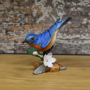 May include: A ceramic figurine of a bluebird perched on a branch with a white flower and green leaves. The bird has blue feathers, orange accents, and a black beak. The figurine is on a brown base.
