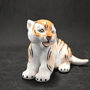 May include: A ceramic figurine of a tiger cub sitting with its paws tucked under its body. The figurine is painted in shades of white, orange, and black to resemble a tiger's stripes. The tiger cub has a playful expression on its face.