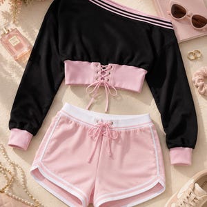 May include: A black and pink two-piece outfit. The off-the-shoulder top has pink stripes and cuffs. The shorts are pink with white trim and a drawstring. A pink book with the text "Je t'aime" is visible.
