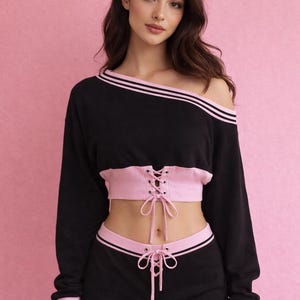 以下が含まれることがあります： 黒とピンクのツーピースアスレチックセット。オフショルダーの長袖トップスはピンクのストライプとトリムが付いています。ショーツはピンクのウエストバンドとトリムが付いています。トップスはコルセットスタイルのレースアップディテールが特徴です。