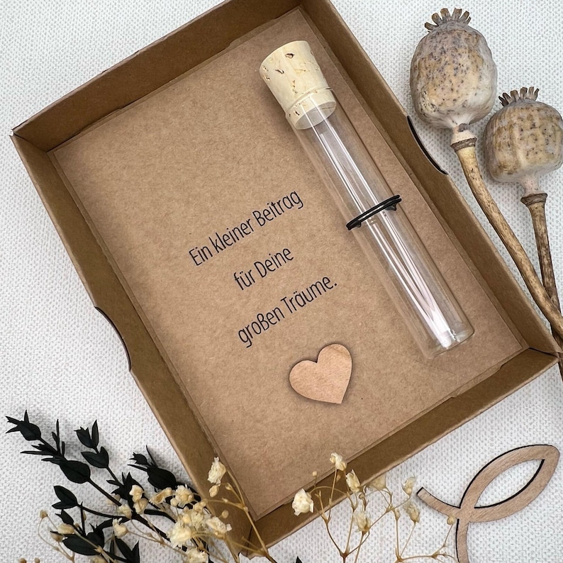 May include: A small brown box with a glass test tube with a cork stopper. The box has a message that reads "Ein kleiner Beitrag f&uuml;r Deine gro&szlig;en Tr&auml;ume." which translates to "A small contribution for your big dreams." There is a small wooden heart in the box.