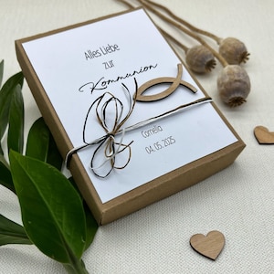 May include: A brown gift box with a white lid tied with twine and a wooden fish charm. The lid has the text "Alles Liebe zur Kommunion" and "Cornelia 04.05.2025" printed on it.