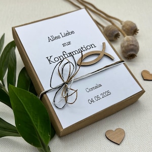 May include: A white gift box with a brown lid tied with a brown, black, and white ribbon. The box has a wooden fish symbol and the text "Alles Liebe zur Konfirmation Cornelia 04.05.2025" printed on it.