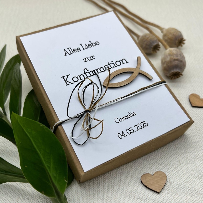May include: A white gift box with a brown lid tied with a brown, black, and white ribbon. The box has a wooden fish symbol and the text "Alles Liebe zur Konfirmation Cornelia 04.05.2025" printed on it.
