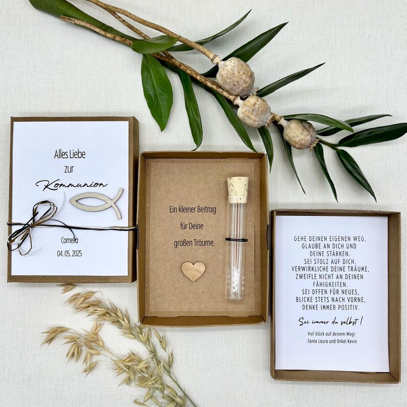 May include: A brown box with a white card inside that says "Alles Liebe zur Kommunion" and "Comella 04.05.2025". A wooden fish is on top of the card. The box also contains a small glass tube with a cork stopper and a wooden heart.
