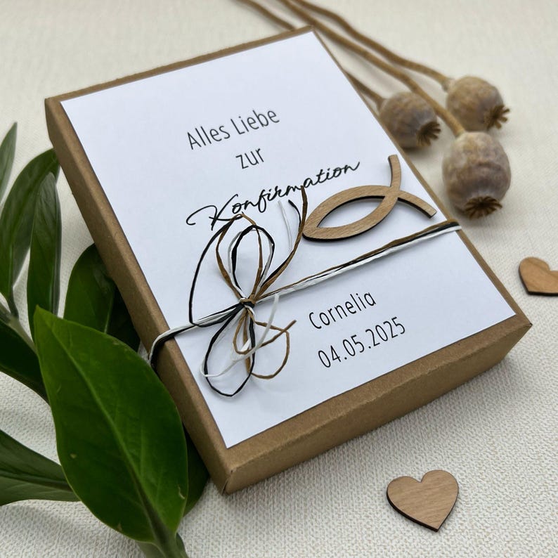 May include: A brown gift box with a white card on top. The card has the text "Alles Liebe zur Konfirmation" and "Cornelia 04.05.2025". A wooden fish is attached to the box with twine.