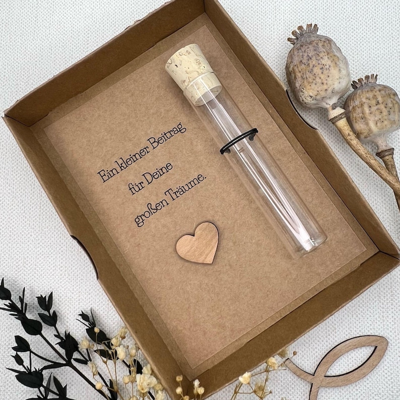 May include: A small brown box with a kraft paper insert. The insert has a message that reads "Ein kleiner Beitrag f&uuml;r Deine gro&szlig;en Tr&auml;ume."  A small wooden heart is attached to the insert. A glass test tube with a cork stopper is placed on the insert.