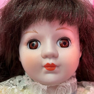 May include: A porcelain doll with brown hair, brown eyes, and red lips. The doll is wearing a white lace dress.