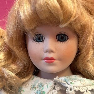 May include: A porcelain doll with blonde hair and blue eyes. The doll has rosy cheeks and red lips. She is wearing a white dress with a floral pattern and a lace collar. The doll's hair is styled in loose curls.