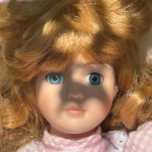 May include: A porcelain doll with light skin, blue eyes, and blonde hair. The doll is wearing a pink and white dress with a lace collar. The doll's hair is styled in loose curls, and it has a delicate, painted face.