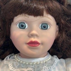 May include: A porcelain doll with brown hair styled in a bun, blue eyes, and red lips. She is wearing a white lace collar with a pearl necklace and a light blue and white striped dress.