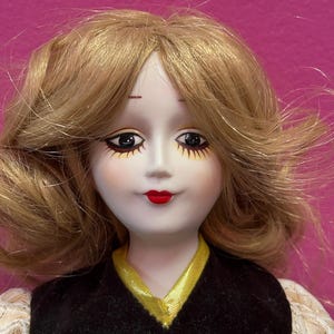 May include: A porcelain doll with blonde hair and a black velvet dress with a gold trim. The doll has red lips, detailed eye makeup, and a pearl button. The doll's dress has lace sleeves.