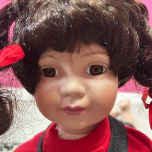 May include: A porcelain doll with brown hair styled in pigtails, adorned with red ribbons. The doll has brown eyes and is wearing a red top with a white collar and black straps. The doll's face is detailed with painted features, including rosy cheeks and a subtle smile.