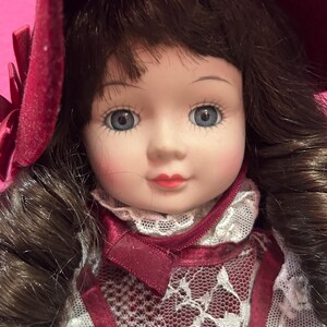 May include: A porcelain doll with blue eyes, rosy cheeks, and red lips. The doll has brown hair and wears a burgundy dress with white lace detailing and a matching hat. The doll's dress has a satin ribbon and a lace collar.