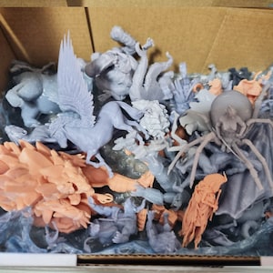 May include: A collection of grey and pink 3D printed miniature figurines, including a winged horse, a spider, and a dragon. The figurines are in a cardboard box.