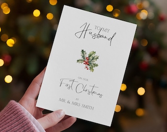 May include: A Christmas card with the message "To My Husband On Our First Christmas" is held in front of a blurred Christmas tree. The card features a holly and berry illustration and the names "Mr & Mrs Smith".