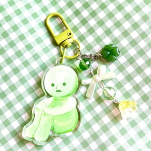 May include: A green and white keychain featuring a seated figure with a sad expression. The keychain has a yellow clasp and is adorned with green beads, a white bow, and a yellow flower charm. The background is a green and white checkered pattern.