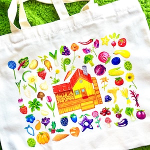 May include: White canvas tote bag featuring a colorful watercolor illustration of a yellow house with a red roof surrounded by various fruits, vegetables, and flowers. The bag has cream-colored handles and is set against a green background.