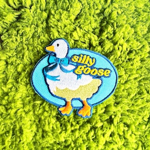 Peut inclure: Écusson brodé représentant une oie de dessin animé avec un nœud bleu et l'inscription "silly goose" en jaune. L'oie a un corps blanc, des pieds jaunes et un contour bleu. L'écusson est posé sur un fond vert vif et texturé.