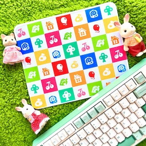 May include: A patterned mat with colourful squares, a mint green keyboard, and three toy rabbits on a bright green, textured surface. The keyboard has white and mint green keys. The mat features houses, leaves, cherries, and balloons.