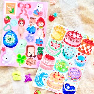 May include: Two sheets of stickers with a pink and blue background. The first sheet features a variety of cute cartoon characters, including a bunny, a squirrel, a fox, and a cat. The second sheet features a variety of colourful cakes with cherries and frosting. The text on the stickers reads "ichigo mort" and "Happy Birthday".