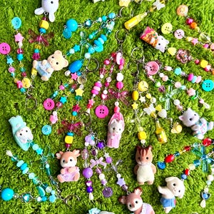 May include: A collection of colorful beaded keychains with various charms, including animal figurines, buttons, and star-shaped beads. The keychains feature a variety of colors, including pink, blue, yellow, and purple, and are arranged on a green, textured surface.