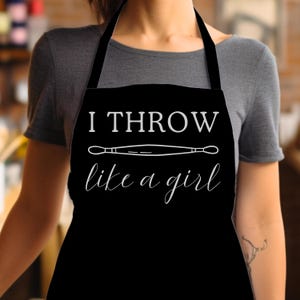 Peut inclure: Tablier noir avec l'inscription blanche "I THROW like a girl". Un dessin au trait blanc d'un rouleau à pâtisserie est centré au-dessus du texte. Le tablier a une sangle de cou noire et des liens.