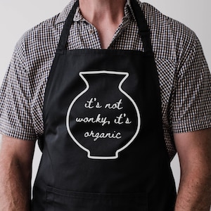 May include: Black apron featuring the text "it's not wonky, it's organic" in white, within a white vase outline. The apron has a bib and neck strap.