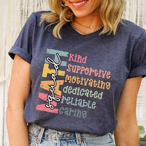 May include: A navy blue t-shirt with a rainbow-colored graphic that reads "Spread Kindness" and lists the words "Kind, Supportive, Motivating, Dedicated, Reliable, Caring".