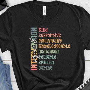 May include: Black t-shirt with a vertical graphic design that reads "Intervention Specialist" with the words "Kind, Supportive, Motivating, Knowledgeable, Dedicated, Reliable, Skilled, Caring" stacked vertically in a rainbow color scheme.