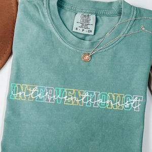 May include: Light green Comfort Colors t-shirt with the word "INTERVENTIONIST" in a colorful, outlined font. A silver necklace with a pendant rests on the shirt. The shirt is folded and has a small tag.