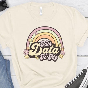 May include: A beige t-shirt with a rainbow and the text "Talk Data To Me" in a retro font. The rainbow is made up of pink, yellow, and orange stripes. There are small yellow flowers around the rainbow.