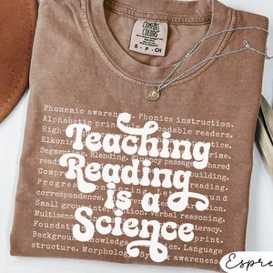 May include: Espresso-colored t-shirt with the text "Teaching Reading is a Science" in white. The shirt has a crew neck and short sleeves. A silver necklace with a pendant is draped across the shirt.
