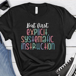 May include: Black t-shirt with the text "But first, explicit, systematic instruction" in colorful letters.
