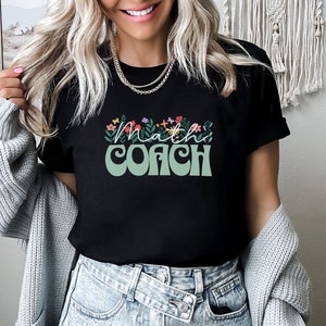 May include: A black t-shirt with the text "Math Coach" in green and white lettering with floral accents. The shirt is being worn by a person with long blonde hair and is paired with light blue jeans.