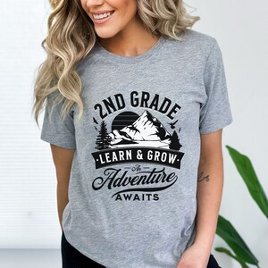 May include: Gray t-shirt with a black graphic design featuring a mountain range, a sunset, and the text "2nd Grade - The Adventure Awaits - Learn & Grow."