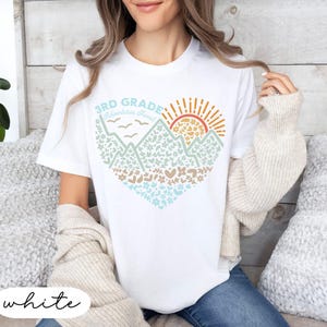 K&ouml;nnte beinhalten: Wei&szlig;es T-Shirt mit einem herzf&ouml;rmigen Berg- und Sonnengrafik. Das Design enth&auml;lt den Text "3rd Grade Adventures Ahead" in Blau und Orange. Die Grafik ist mit Blumen- und Herzmustern gef&uuml;llt.