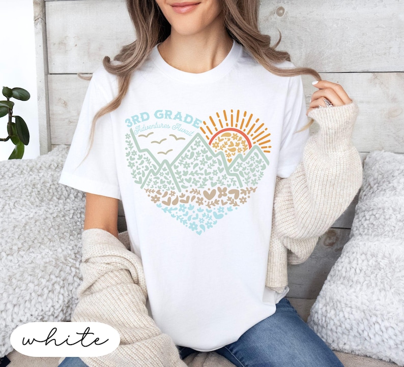 K&ouml;nnte beinhalten: Wei&szlig;es T-Shirt mit einem herzf&ouml;rmigen Berg- und Sonnengrafik. Das Design enth&auml;lt den Text "3rd Grade Adventures Ahead" in Blau und Orange. Die Grafik ist mit Blumen- und Herzmustern gef&uuml;llt.