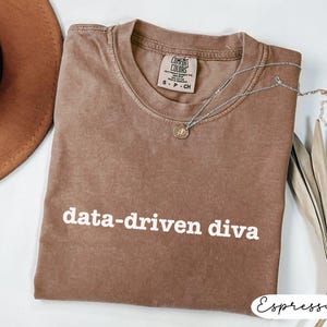 May include: Espresso-colored t-shirt with the phrase "data-driven diva" in white block letters. A silver necklace with a small pendant rests on the shirt. The shirt is a Comfort Colors brand, size S-P-CH.