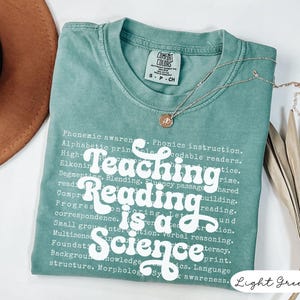 May include: Light green t-shirt with the text "Teaching Reading is a Science" in white. The shirt has a crew neck and short sleeves. A silver necklace with a pendant is draped across the shirt.