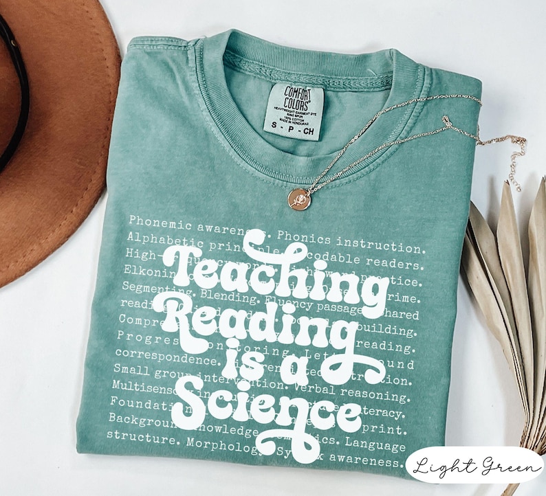May include: Light green t-shirt with the text "Teaching Reading is a Science" in white. The shirt has a crew neck and short sleeves. A silver necklace with a pendant is draped across the shirt.