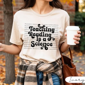 May include: Ivory-colored t-shirt with the text "Teaching Reading is a Science" in black script. The shirt is worn with a plaid scarf and blue jeans. A white coffee cup is held in the hand.