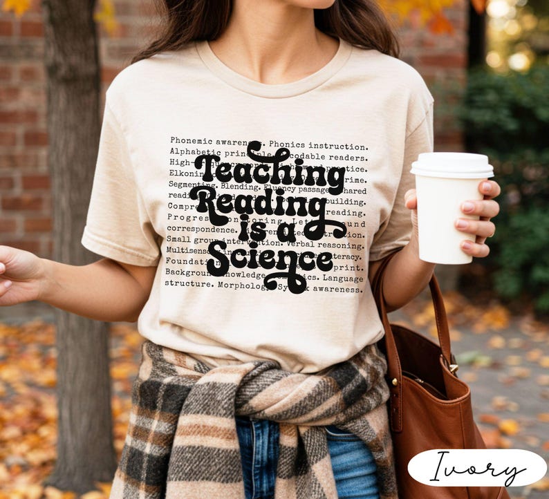 May include: Ivory-colored t-shirt with the text "Teaching Reading is a Science" in black script. The shirt is worn with a plaid scarf and blue jeans. A white coffee cup is held in the hand.