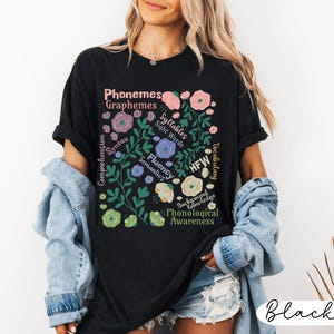 May include: Black t-shirt with a floral design and text related to phonological awareness. The design includes words like "Phonemes," "Graphemes," "Syllables," and "Sight Words" in a floral arrangement. The shirt is a casual wear item.