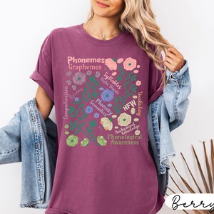 May include: Berry-colored t-shirt with a floral design and educational terms like "Phonemes," "Graphemes," and "Syllables." The design includes flowers and leaves in various colors. The shirt has short sleeves and a relaxed fit.