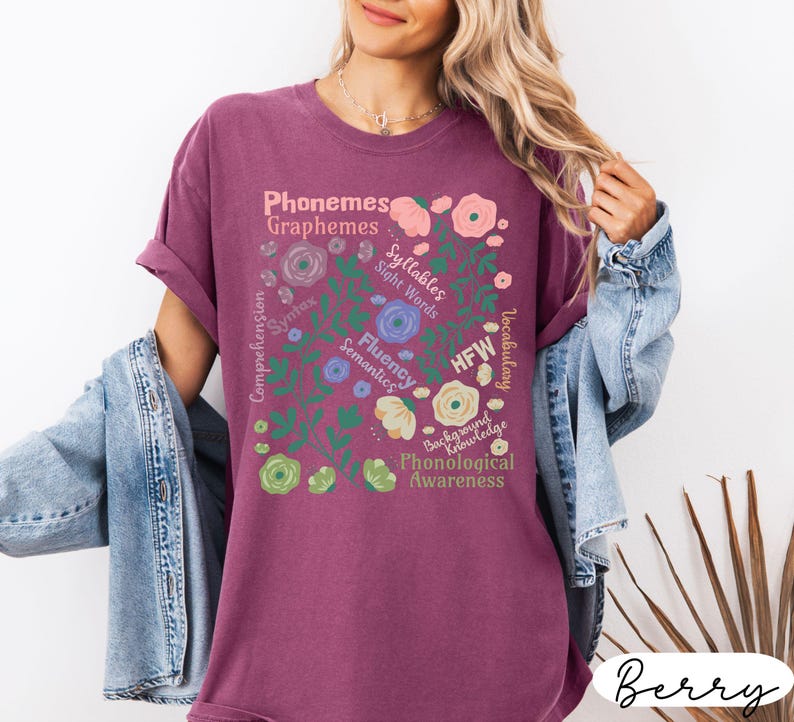 May include: Berry-colored t-shirt with a floral design and educational terms like "Phonemes," "Graphemes," and "Syllables." The design includes flowers and leaves in various colors. The shirt has short sleeves and a relaxed fit.